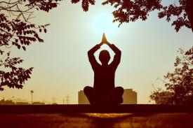 Person in nature sitting with their legs crossed, arms up above their head with palms touching each other